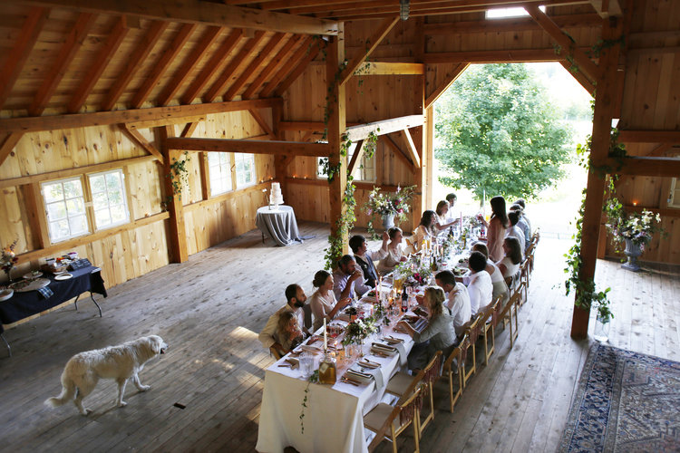 People eating at long family table set with white linen inside rustic barn, with a white dog