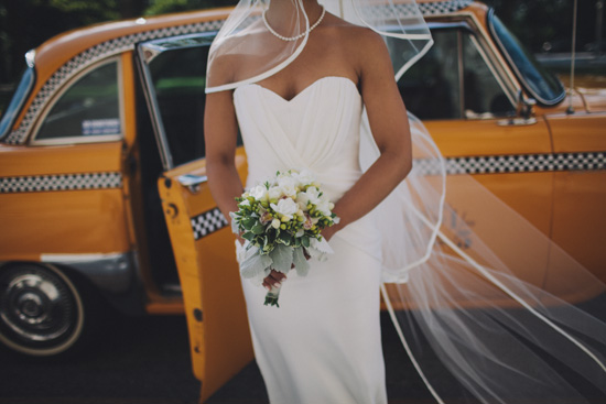 Woman in white strapless wedding dress with veil holding bouquet in front of a yellow checkered cab