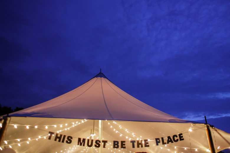 Party tent illuminated with white string lights with a banner that reads "This must be the place"