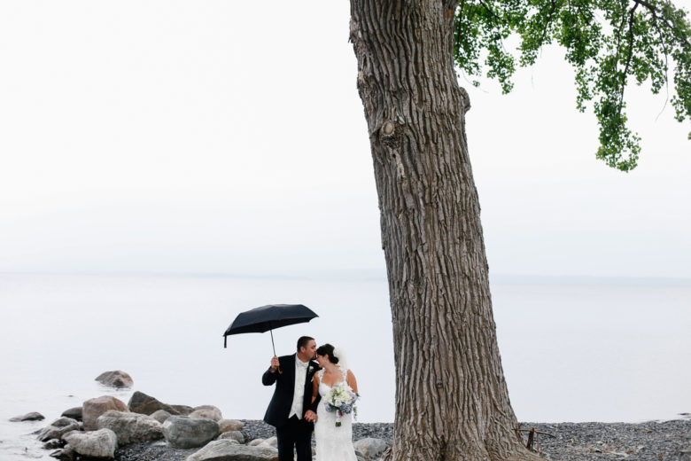 Newly wed couple under giant tree in front of water with large rocks, with groom holding black umbrella overhead