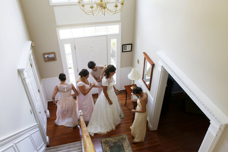 Two women fastening bride's dress downstairs in house foyer, with two other women