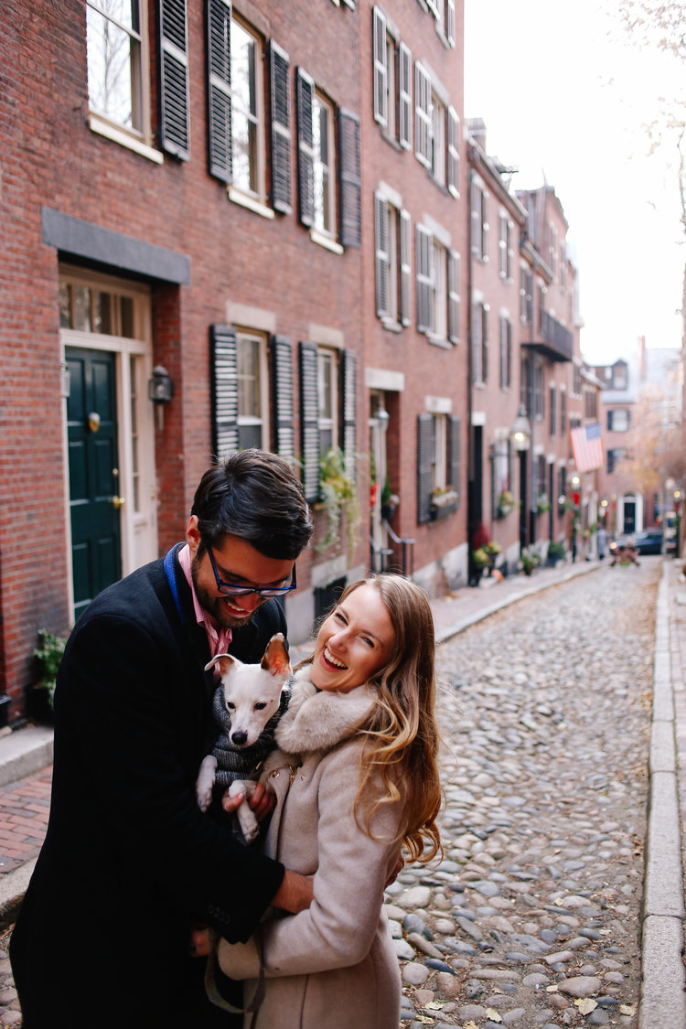 Smiling couple on cobblestone street with dog wearing a puffy vest