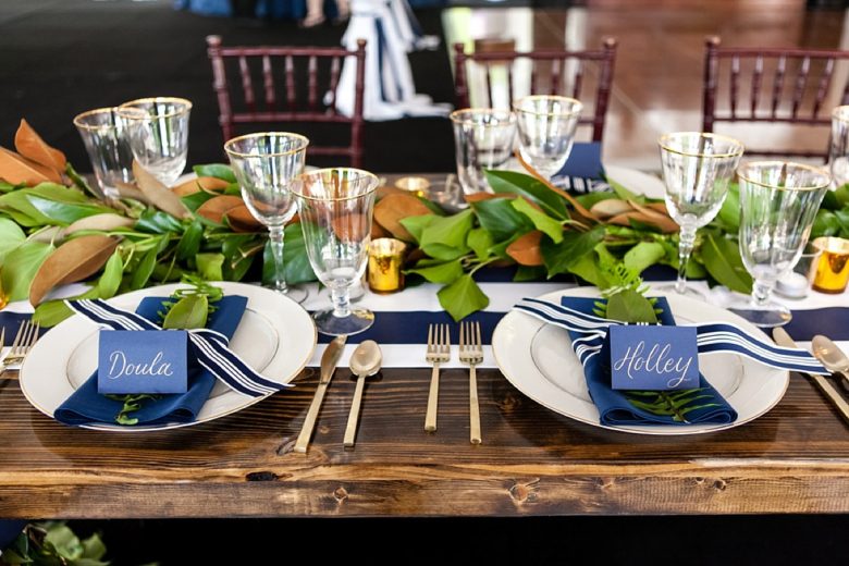 preppy tablescape with blue napkins, a blue and white striped table runner, leafy greenery along runner, glass goblets, gold utensils, on polished wood table, with blue place cards