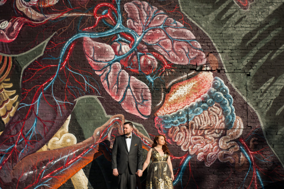 man in suit and woman in gold dress hold hands in front of wall with anatomical heart mural