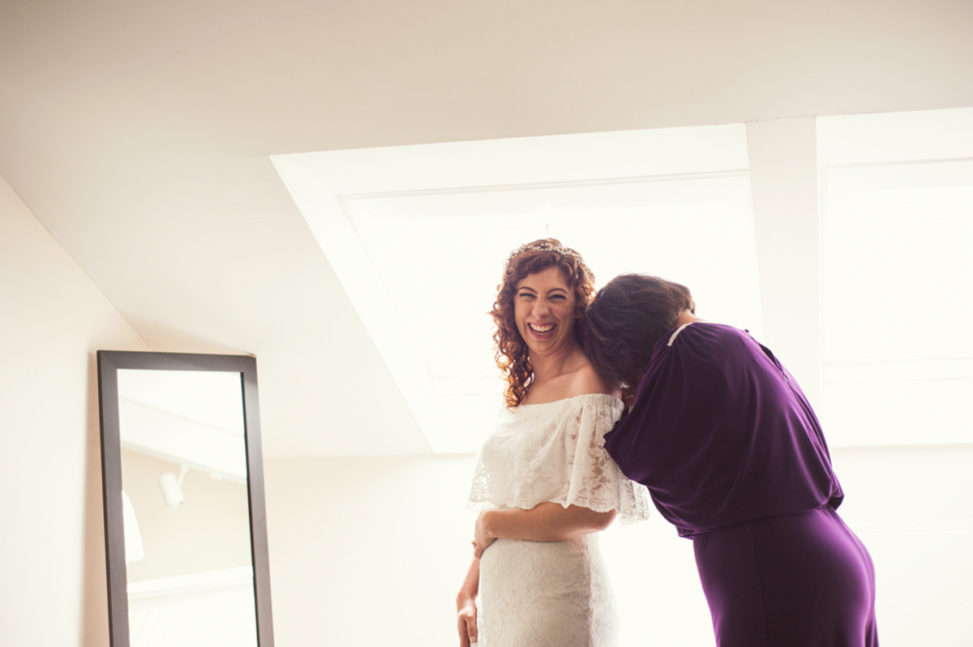 woman in off the shoulder wedding gown looking at camera and smiling while bridesmaid buries her head laughing