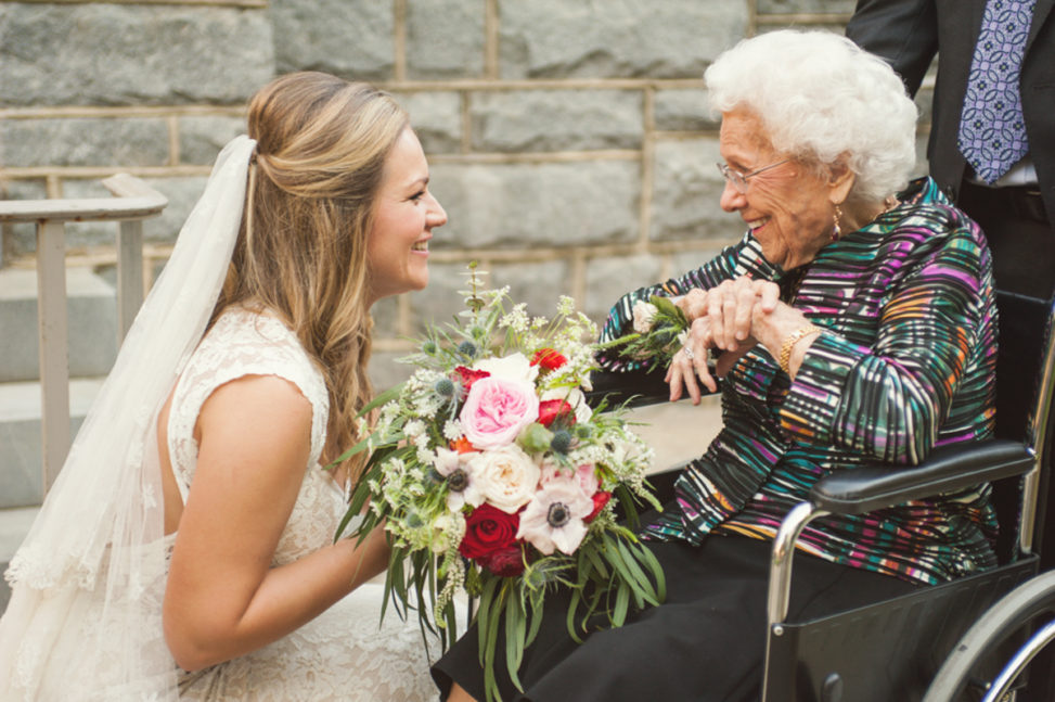 bride holding bouquet kneels in front of elderly lady in wheelchair