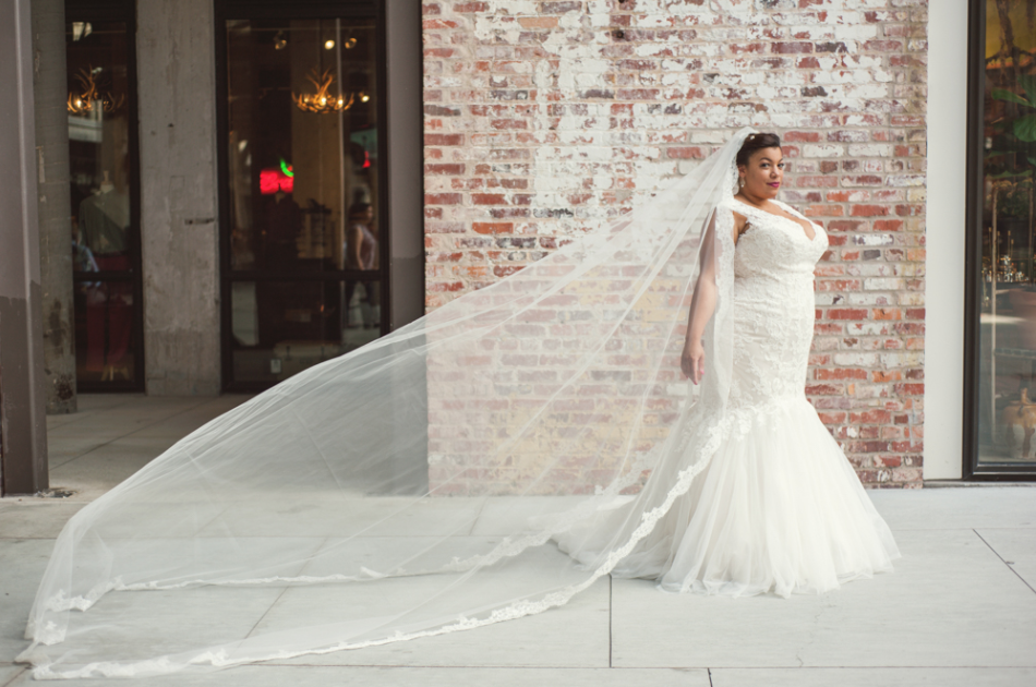 plus size black bride in front of a brick wall in a mermaid wedding gown and cathedral length veil blowing behind her